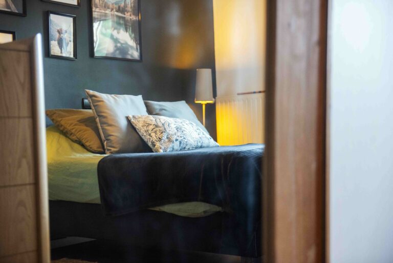 Ambiance cosy du projet Dressing chic par Bulle Intérieure : lit habillé de coussins, mur foncé décoré de cadres et lumière tamisée d’une lampe