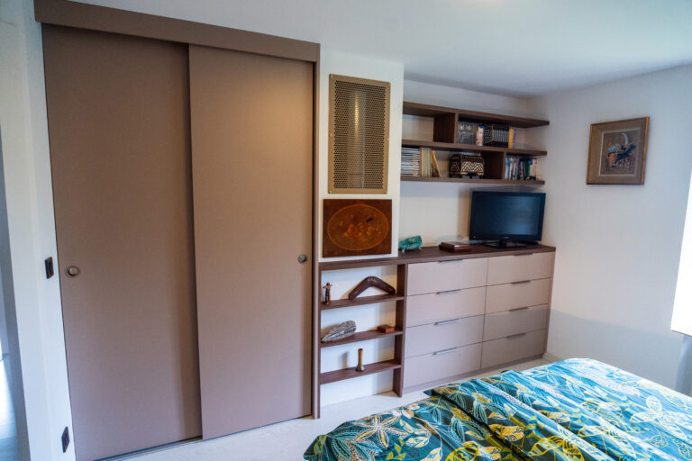 Mobilier sur-mesure dans une chambre rénovée, avec dressing à portes coulissantes, commode intégrée et grille en métal perforé pour le conduit de cheminée.