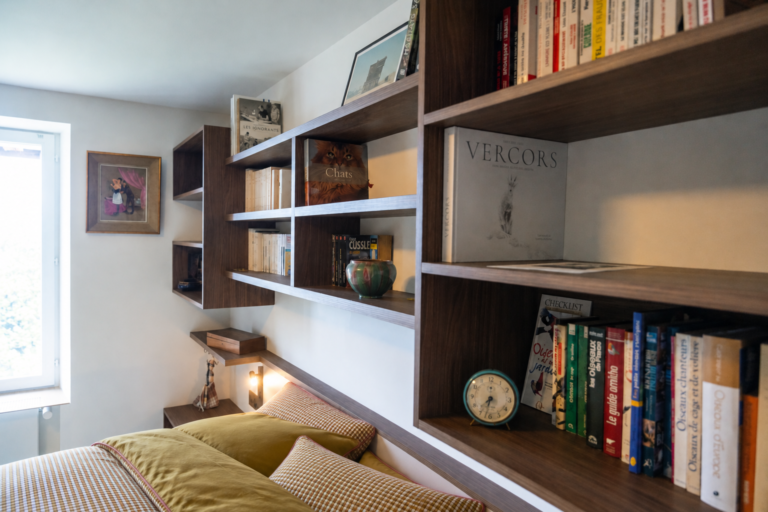 Vue rapprochée d'une bibliothèque suspendue en bois foncé, avec niches ouvertes et étagères intégrées au-dessus du lit dans une chambre du Vercors.
