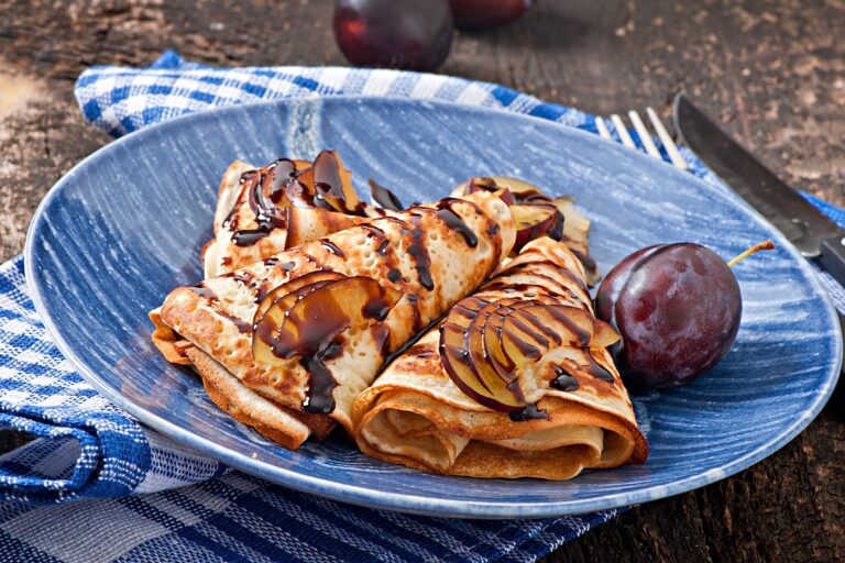 Crêpes bretonnes pliées avec prunes fraîches et nappage au chocolat sur une assiette bleue.